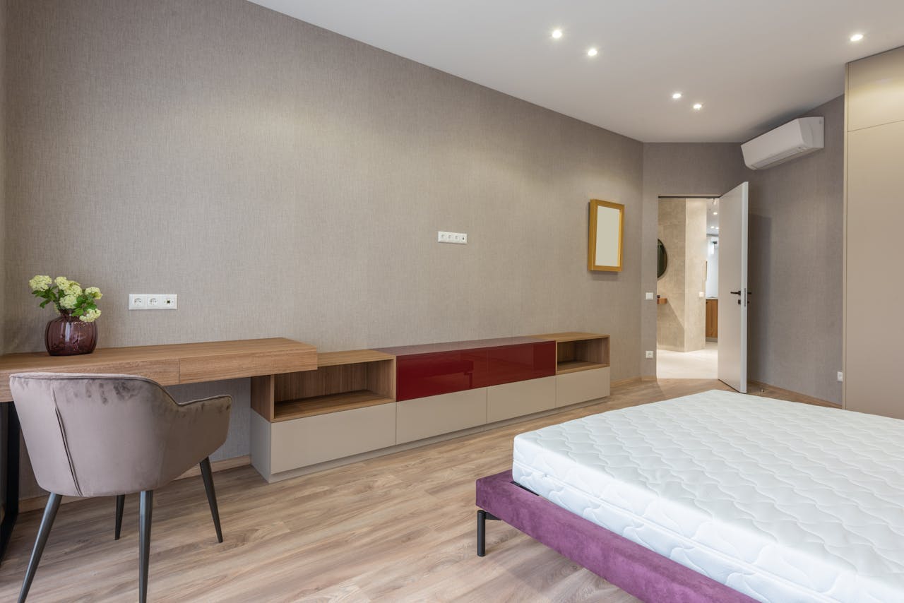 Soft bed with mattress placed against wooden table with decorative flowerpot in spacious stylish bedroom with opened door at home